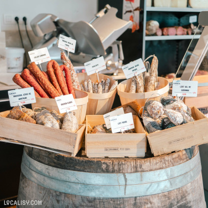 Un étal de charcuterie dans la boucherie Simonis Boucherie Traiteur à Plainevaux. Plusieurs types de saucissons sont disposés dans des paniers et des caisses en bois sur un tonneau. Chaque type de saucisson est étiqueté avec son nom et son prix.