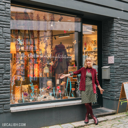 Devanture du magasin d’art et d’artisanat Il était une fée à Huy, la vitrine est ornée de divers objets et décorations, avec un texte en français sur la vitre qui dit : Chez H, enfant une fée, on récolte ce que les enfants sèment. Une personne se tient debout devant le magasin créant une ambiance chaleureuse et accueillante.