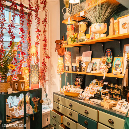 Des étagères remplies d'objets divers tels que des cadres photo, des bijoux, des lampes, des vases et des articles de papeterie dans le magasin d’art et d’artisanat Il était une fée à Huy. Des guirlandes de fleurs rouges pendent du plafond, ajoutant une touche colorée et festive à l'espace.