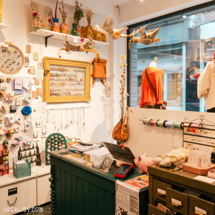 L'intérieur du magasin d’art et d’artisanat Il était une fée à Huy. On y voit un comptoir avec divers objets, notamment des bijoux, des accessoires, et des fournitures d'artisanat. Il y a des étagères avec des vases colorés et des coussins décoratifs, ainsi que des oiseaux suspendus au plafond.