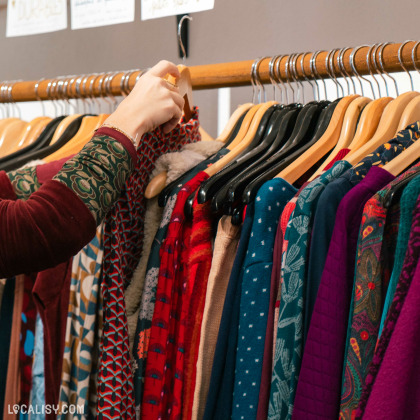 Une personne en train de choisir des vêtements sur un portant dans le magasin d’art et d’artisanat Il était une fée à Huy. Le portant contient une variété de vêtements colorés, principalement des hauts avec différents motifs et textures.