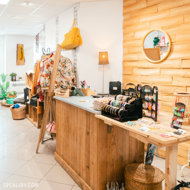 Un comptoir en bois avec des présentoirs de bijoux, des vêtements suspendus sur des étagères, et divers objets de décoration à l'intérieur du magasin de vêtements Annestramgram à Aywaille. Le mur à droite est recouvert de panneaux de bois, et il y a un miroir rond accroché dessus.