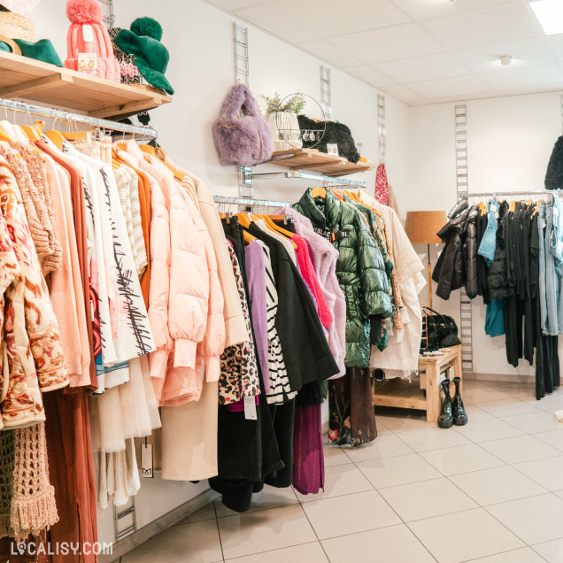 Plusieurs portants avec des vêtements suspendus, notamment des manteaux, des vestes, des pulls et des robes de différentes couleurs et styles au magasin de vêtements Annestramgram à Aywaille. Sur les étagères au-dessus des portants, il y a des accessoires comme des chapeaux, des sacs et des écharpes.