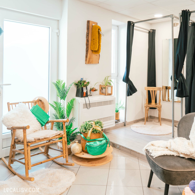 Un espace intérieur confortable et bien décoré du magasin de vêtements Annestramgram à Aywaille. Il y a une chaise berçante en bois avec un coussin blanc moelleux et un sac vert accroché dessus. À côté de la chaise se trouve une table ronde en bois avec un sac à main vert et un panier de fleurs.