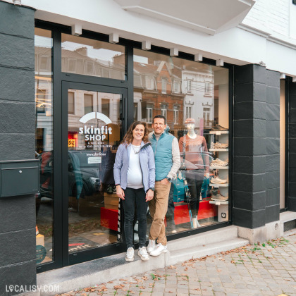 Devanture du magasin de vêtements de sport Skinfit Shop Verviers à Heusy. La vitrine du magasin affiche des vêtements et des chaussures. Les heures d'ouverture sont indiquées sur la porte : Mo 14:00-18:00, Tu-Fr 10:00-18:00, Sa 10:00-17:00. Deux personnes se tiennent debout devant l'entrée du magasin.