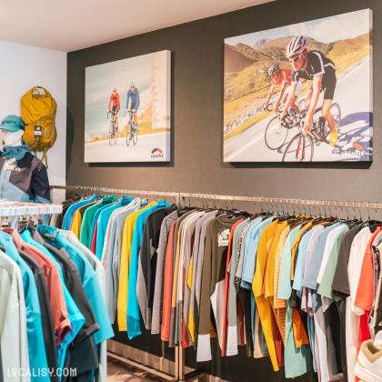 L'intérieur du magasin de vêtements de sport Skinfit Shop Verviers à Heusy avec des portants remplis de chemises et de vestes colorées. Au-dessus des portants, il y a deux grandes affiches représentant des cyclistes sur des routes, avec le nom de la marque Skinfit visible sur les affiches. À gauche, un sac à dos jaune est accroché au mur.