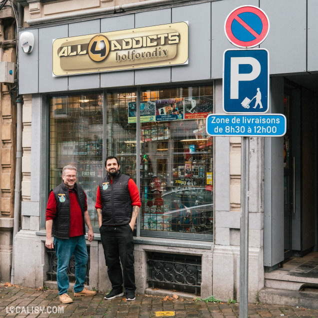 Devanture du magasin de jeux vidéo All4Addicts à Verviers. Deux personnes se tiennent devant le magasin, portant des gilets noirs et des vêtements rouges. La vitrine du magasin expose divers produits, notamment des jeux et des figurines. À droite, il y a un panneau de signalisation indiquant une zone de livraisons de 8h30 à 12h00.