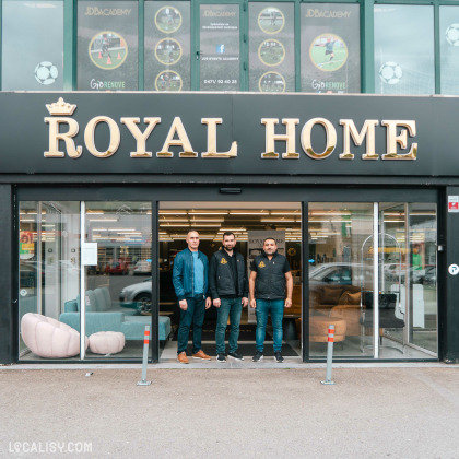 Devanture du magasin de Meubles & Accessoires Royal Home à Verviers. Trois personnes se tiennent devant l'entrée du magasin, qui a une grande enseigne noire avec des lettres dorées indiquant ROYAL HOME. À l'intérieur, on peut voir des meubles, notamment un canapé bleu et un fauteuil rose. Au-dessus de l'enseigne, il y a des affiches pour JDB Academy et GioRenove.