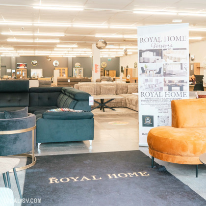 L'intérieur du magasin de Meubles & Accessoires Royal Home à Verviers. On y voit plusieurs canapés et fauteuils exposés, avec un grand tapis noir portant l'inscription ROYAL HOME au premier plan. Un panneau publicitaire à droite affiche des informations sur le magasin, y compris des photos de meubles et des détails de contact.