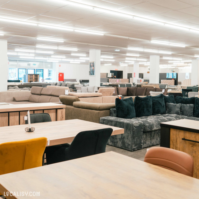 Une grande variété de canapés et de fauteuils disposés dans un espace bien éclairé à l'intérieur du magasin de Meubles & Accessoires Royal Home à Verviers. À l'avant-plan, il y a des tables en bois avec des chaises de différentes couleurs.