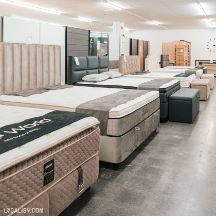 Une rangée de matelas exposés dans le magasin de Meubles & Accessoires Royal Home à Verviers. Chaque matelas est accompagné d'une tête de lit et certains ont des oreillers posés dessus. Les matelas sont de différentes marques et modèles, et sont disposés sur des sommiers. Le sol est en carrelage gris et l'éclairage est fluorescent. Il y a également des meubles en bois à l'arrière-plan.