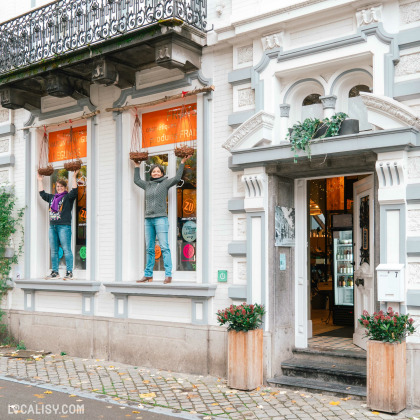 Devanture de l'épicerie Demain l'Épicerie à Spa. Deux personnes sont debout sur les rebords des fenêtres, tenant des paniers suspendus. Les fenêtres ont des autocollants colorés et des inscriptions en français. L'entrée du magasin est ouverte, révélant l'intérieur avec des étagères et des produits.