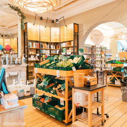 Des étagères en bois garnies de produits locaux et naturels dans l'épicerie Demain l'Épicerie à Spa. On y voit des légumes frais dans des cagettes vertes, des distributeurs en vrac, ainsi que des étagères remplies de produits variés dans des contenants réutilisables.