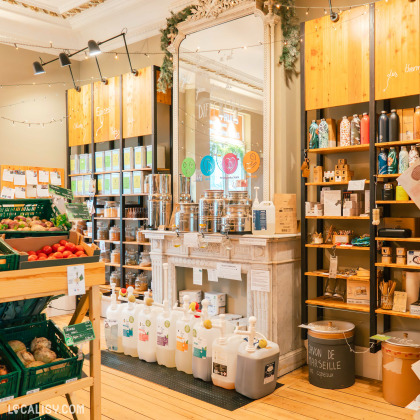 Des étagères en bois remplies de produits divers, des distributeurs de liquides en vrac sur une cheminée décorative, et des paniers de fruits et légumes frais à l'intérieur de l'épicerie Demain l'Épicerie à Spa. L'ambiance est chaleureuse avec des guirlandes lumineuses et une décoration soignée.