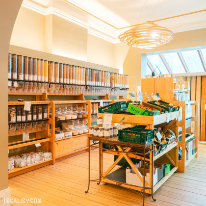 L'intérieur de l'épicerie Demain l'Épicerie à Spa, avec des distributeurs en vrac alignés sur les murs, contenant divers produits secs comme des céréales, des légumineuses et des noix. Au centre, il y a une table avec des caisses de fruits et légumes frais, notamment des kiwis.