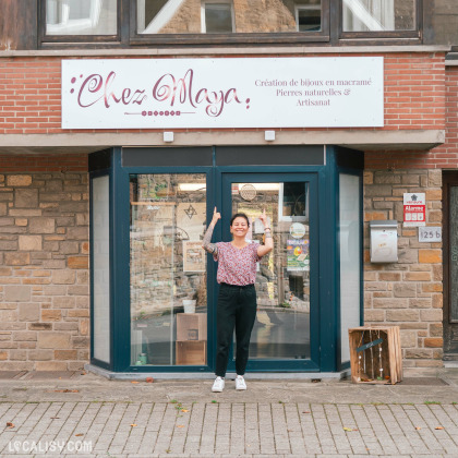 Devanture du magasin d'artisanat Chez Maya à Tilff. Le magasin a une enseigne blanche avec le texte Chez Maya en lettres cursives rouges et Création de bijoux en macramé, Pierres naturelles & Artisanat en lettres plus petites. La façade est en pierre et brique, avec une grande porte vitrée bleue. Une personne se tient devant le magasin, pointant vers l'enseigne.