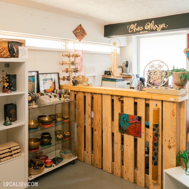 L'intérieur du magasin d'artisanat Chez Maya à Tilff, avec un comptoir en bois fait de palettes, des étagères avec des bols chantants, des bracelets, des œuvres d'art encadrées, des attrape-rêves, et d'autres objets décoratifs. Il y a également des plantes et des objets en bois sculpté.