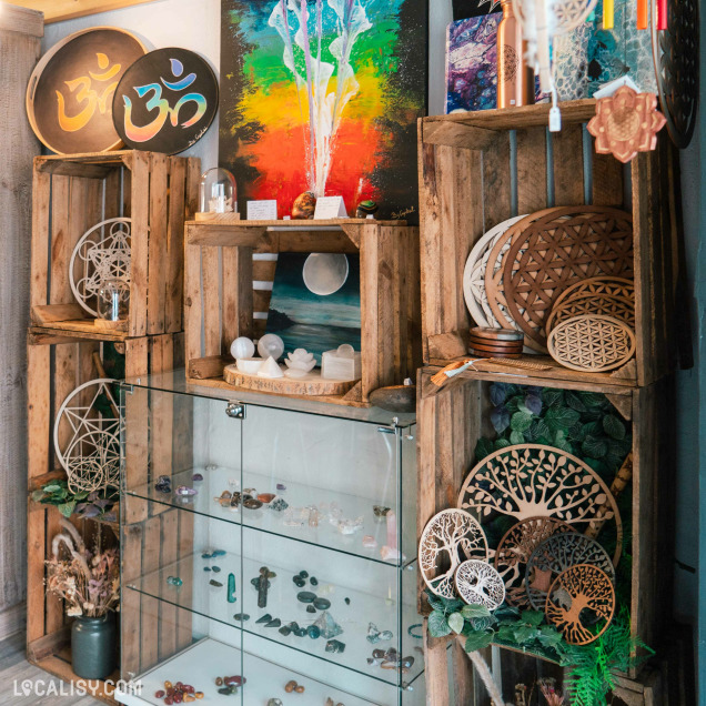 Un présentoir composé de caisses en bois empilées, contenant divers objets décoratifs et spirituels dans le magasin d'artisanat Chez Maya à Tilff. On y voit des disques en bois avec des motifs géométriques et des arbres de vie, des pierres et cristaux exposés dans une vitrine en verre, ainsi que des œuvres d'art colorées accrochées au mur.