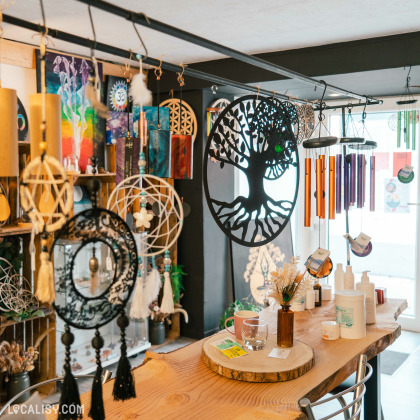 Une variété de décorations suspendues, y compris des attrape-rêves, des carillons éoliens, et des objets en forme d'arbre de vie dans le magasin d'artisanat Chez Maya à Tilff. Il y a également des étagères avec des œuvres d'art colorées et des produits divers comme des bougies et des crèmes. La table en bois au centre est décorée avec des articles de bien-être et des échantillons de produits.