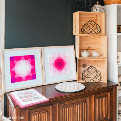 Un coin du magasin d'artisanat Chez Maya à Tilff avec des éléments de décoration. Il y a deux cadres avec des motifs géométriques colorés, principalement en rose et violet, posés sur un meuble en bois. À côté, il y a des étagères en bois contenant des objets décoratifs, dont des structures métalliques géométriques et des petits bols. Sur le meuble, il y a également un livre ouvert et un plateau rond avec un motif géométrique.