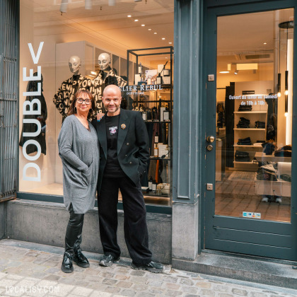 Devanture du magasin de vêtements pour femmes Double V à Liège, avec une vitrine qui expose des vêtements et une enseigne indique ATELIER REBUL DEPUIS 1895. Deux personnes debout devant le magasin. La porte affiche les horaires d'ouverture : Ouvert du lundi au samedi de 10h à 18h.