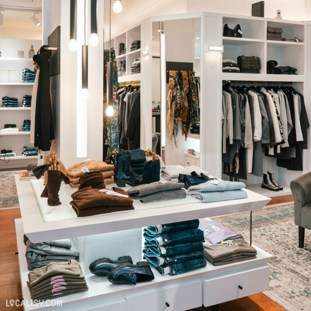 L'intérieur du magasin de vêtements pour femmes Double V à Liège, avec des étagères et des portants remplis de vêtements soigneusement pliés et suspendus. Il y a des pulls, des pantalons, des vestes et des accessoires comme des chaussures et des sacs.