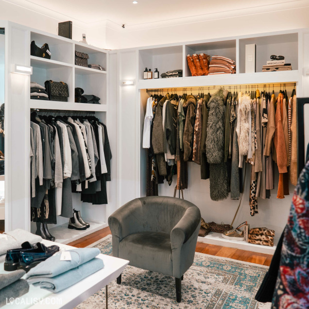 Un espace bien organisé avec des vêtements suspendus sur des cintres et des étagères contenant des sacs à main et des accessoires dans le magasin de vêtements pour femmes Double V à Liège. Il y a également un fauteuil gris au centre de la pièce et une table avec des vêtements pliés.