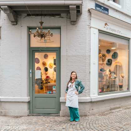 Devanture de la Chapellerie Le Chapeau d'Or à Liège. Une personne portant un tablier blanc se tient devant la porte d'entrée du magasin. La vitrine expose plusieurs chapeaux de différentes couleurs et styles. Une enseigne au-dessus de la vitrine indique le nom du magasin. Le magasin est situé à l'angle d'une rue pavée, avec un panneau de rue visible indiquant Rue Chapallée des Clercs.