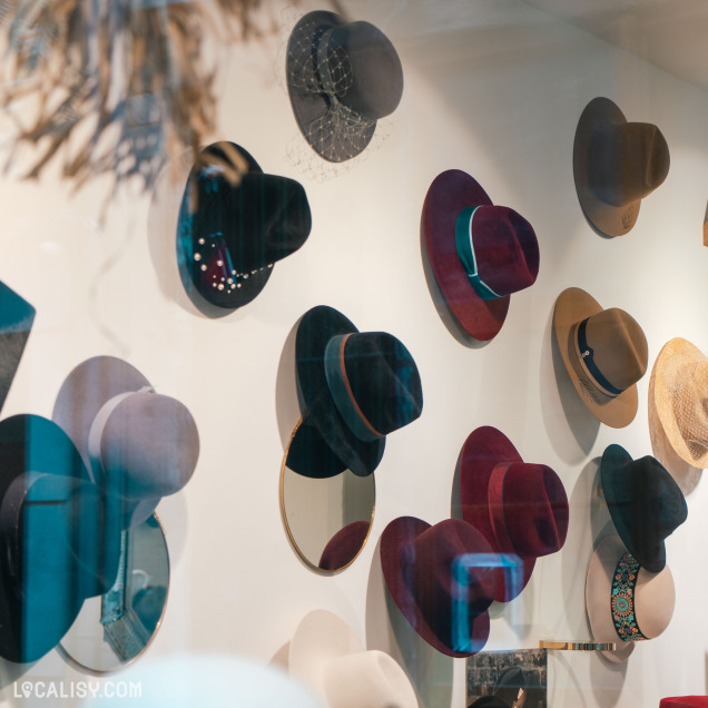 Un mur d'exposition dans la Chapellerie Le Chapeau d'Or à Liège, avec plusieurs chapeaux de différentes couleurs et styles accrochés. Les chapeaux sont disposés de manière artistique, certains étant placés devant des miroirs ronds.