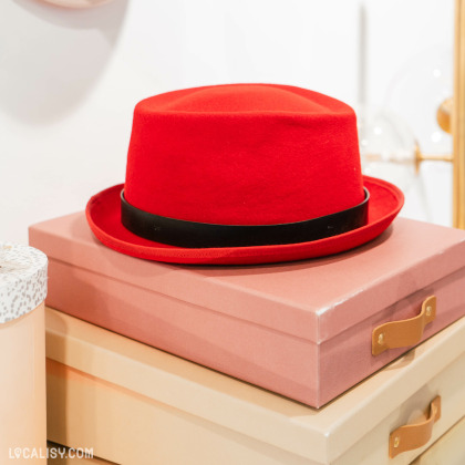 Un chapeau rouge vif avec un ruban noir posé sur une boîte rose. Le chapeau a une forme classique de fedora. La boîte rose est placée sur une autre boîte beige avec une poignée en cuir dans la Chapellerie Le Chapeau d'Or à Liège.