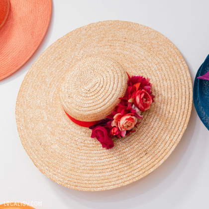 Un chapeau de paille beige orné de fleurs rouges et roses sur le côté, avec un ruban rouge à l'intérieur de la Chapellerie Le Chapeau d'Or à Liège. Ce chapeau est entouré par deux autres chapeaux, un orange à gauche et un bleu à droite.