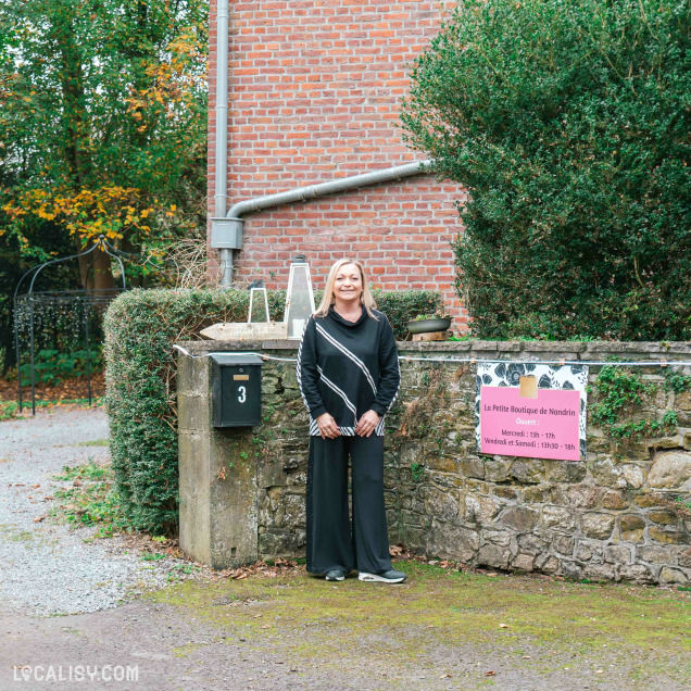 Devanture du magasin de vêtements pour femmes La Petite Boutique de Nandrin à Nandrin. Une personne debout devant un mur en pierre avec une boîte aux lettres noire marquée du numéro 3. À droite de la personne, il y a une affiche rose indiquant les horaires d'ouverture de La Petite Boutique de Nandrin. L'affiche indique : La Petite Boutique de Nandrin Ouvert: Mercredi: 13h - 17h Vendredi et Samedi: 13h30 - 18h. En arrière-plan, on peut voir une maison en briques rouges et de la végétation.