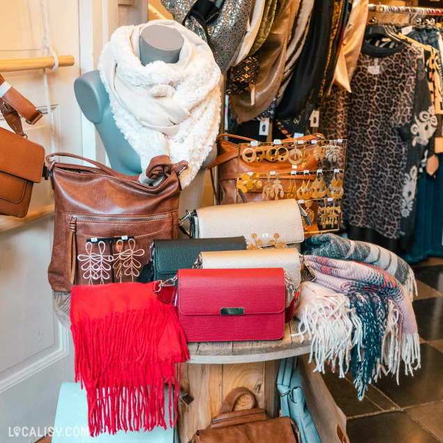 Un présentoir dans le magasin de vêtements pour femmes La Petite Boutique de Nandrin à Nandrin, avec divers articles de mode, notamment des sacs à main en cuir de différentes couleurs, des écharpes, des foulards, et des bijoux comme des boucles d'oreilles. Un mannequin porte une écharpe blanche en laine.