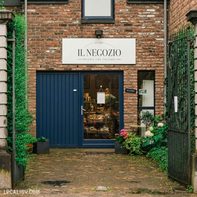 Devanture de l'épicerie Il Negozio à Verviers. La façade est en briques avec une porte bleue et une grande fenêtre. Au-dessus de la porte, il y a une enseigne blanche avec le texte IL NEGOZIO et EPICERIE FINE ITALIENNE. Devant le magasin, il y a des plantes en pot et des fleurs. L'intérieur du magasin est visible à travers la fenêtre, montrant des étagères avec des produits.