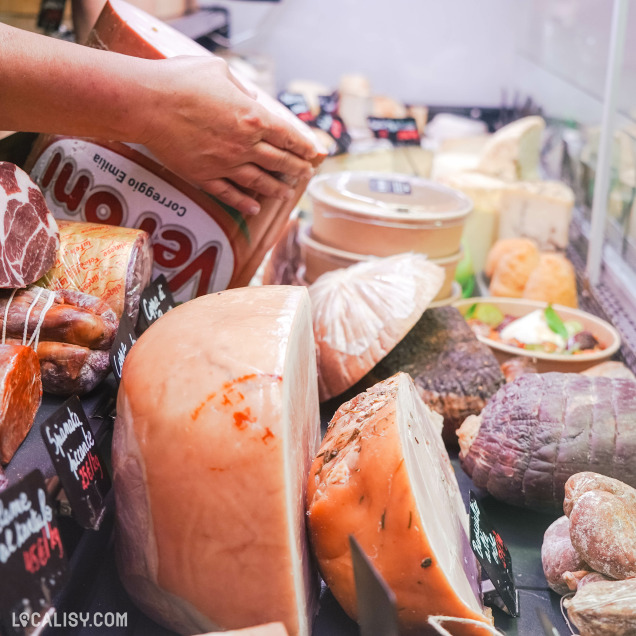 Un étal de charcuterie dans l'épicerie Il Negozio à Verviers, avec une variété de produits de charcuterie, y compris des jambons, des saucisses, et d'autres viandes séchées et fumées. Les produits sont étiquetés avec des petites ardoises indiquant les noms et les prix. Une personne est en train de manipuler un gros morceau de viande emballé.