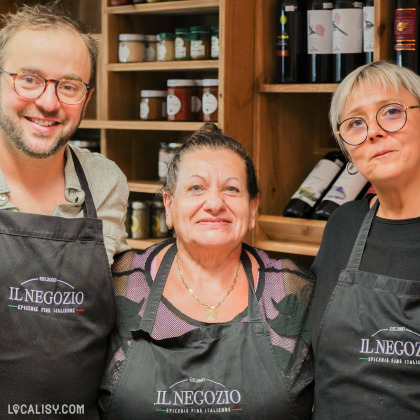 Trois personnes portant des tabliers noirs avec le logo IL NEGOZIO ÉPICERIE FINE ITALIENNE brodé dessus à l'épicerie Il Negozio à Verviers. Elles se tiennent devant des étagères en bois remplies de produits alimentaires, notamment des pots de confiture et des bouteilles de vin.