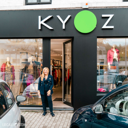 Devanture du magasin de vêtements pour femmes Kyoz à Tilff. Le nom du magasin est affiché en grandes lettres blanches sur un fond noir, avec un cercle vert remplaçant le O. Devant le magasin, une personne se tient debout sur le trottoir. À l'intérieur de la vitrine, on peut voir des mannequins habillés avec des vêtements colorés. Des voitures sont garées de part et d'autre de l'entrée du magasin.