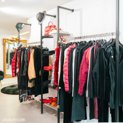 L'intérieur du magasin de vêtements pour femmes Kyoz à Tilff. On y voit des portants avec divers vêtements suspendus, principalement dans des tons de rouge, noir et doré. Il y a également des accessoires comme des chapeaux et des sacs à main exposés sur les étagères au-dessus des portants. Un grand miroir doré est visible en arrière-plan.