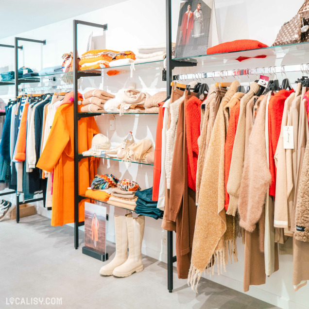 L'intérieur du magasin de vêtements pour femmes Kyoz à Tilff, avec des étagères et des portants remplis de vêtements colorés et divers accessoires. On y voit des manteaux, des pulls, des écharpes, des chaussures et des bottes, principalement dans des tons chauds comme l'orange, le beige et le marron.