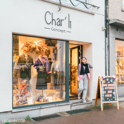 Devanture du magasin de vêtements Char'li Concept à Malmedy. La vitrine présente des vêtements et des accessoires, avec une décoration florale suspendue. Une personne se tient à l'entrée du magasin. À droite de l'entrée, un panneau indique NOUS SOMMES OUVERTS WE ARE OPEN.