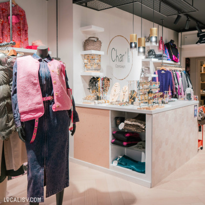 L'intérieur du magasin de vêtements Char'li Concept à Malmedy, avec un mannequin vêtu d'une robe en jean et d'un gilet rose, placé à côté d'un comptoir. Le comptoir est décoré avec divers bijoux et accessoires exposés. Derrière le comptoir, il y a des étagères avec des sacs à main et des vêtements suspendus.