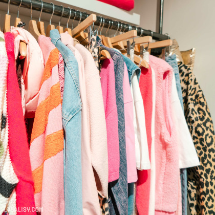 Une rangée de vêtements suspendus sur des cintres dans le magasin de vêtements Char'li Concept à Malmedy. Les vêtements sont principalement dans des tons de rose, rouge, et bleu, avec des motifs variés, y compris des rayures et des imprimés léopard.