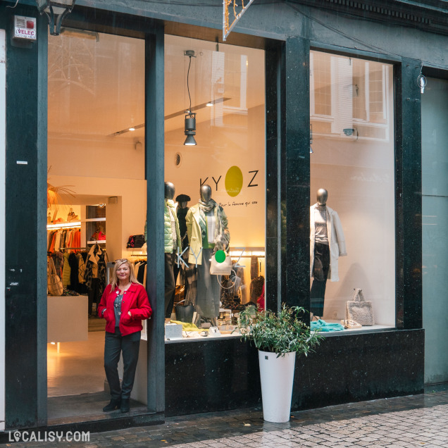 Devanture du magasin de vêtements pour femmes Kyoz à Liège. La vitrine présente plusieurs mannequins habillés avec des vêtements modernes et élégants. Une personne portant une veste rouge se tient à l'entrée du magasin. Le logo du magasin Kyoz est visible sur la vitre, accompagné du slogan : Pour la femme que vous êtes.