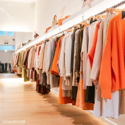 Une rangée de vêtements suspendus sur des cintres le long d'une étagère éclairée dans le magasin de vêtements pour femmes Kyoz à Liège. Les vêtements sont variés en termes de couleurs et de styles, avec une prédominance de tons orange et neutres. L'éclairage est chaleureux et met en valeur les articles exposés.