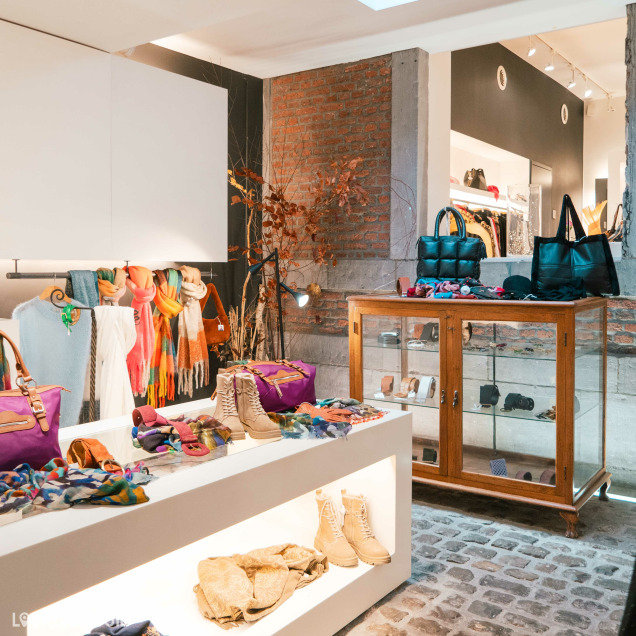 Des écharpes colorées accrochées à des cintres, des sacs à main, des bottes et divers accessoires disposés sur des présentoirs et dans une vitrine en bois au magasin de vêtements pour femmes Kyoz à Liège. .Le magasin a une ambiance chaleureuse avec des murs en briques apparentes et un éclairage doux.