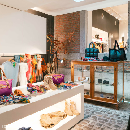 Des écharpes colorées accrochées à des cintres, des sacs à main, des bottes et divers accessoires disposés sur des présentoirs et dans une vitrine en bois au magasin de vêtements pour femmes Kyoz à Liège. .Le magasin a une ambiance chaleureuse avec des murs en briques apparentes et un éclairage doux.