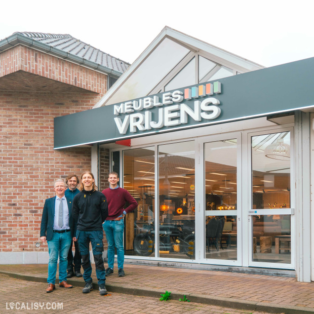 Devanture du magasin de meubles Vrijens à Juprelle. On peut voir quatre personnes debout devant le magasin. L'enseigne du magasin affiche MEUBLES VRIJENS avec un logo coloré à côté. Le bâtiment est en briques avec un toit incliné, et l'intérieur du magasin est visible à travers les grandes fenêtres vitrées.