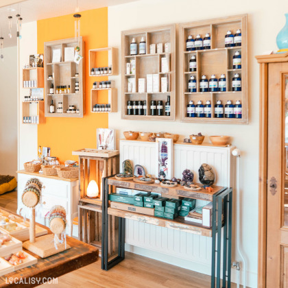 L'intérieur du magasin de minéraux Alma Nature à Hamoir, avec des étagères en bois accrochées au mur, contenant des bouteilles et des boîtes de produits divers. Sur les tables et les étagères, il y a des pierres précieuses, des cristaux, et des objets décoratifs. Il y a également des paniers en osier contenant des articles à vendre.