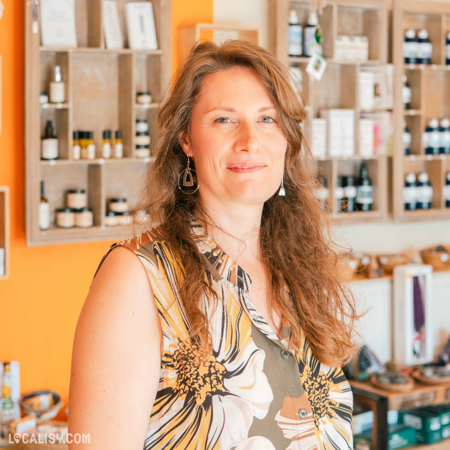 L'intérieur du magasin de minéraux Alma Nature à Hamoir. On peut voir des étagères en bois fixées au mur, contenant divers produits, notamment des bouteilles et des pots. Au premier plan, une personne porte un haut à motifs floraux.