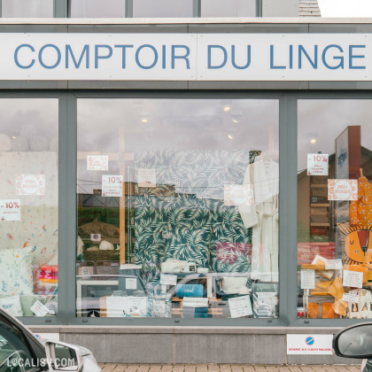 Devanture du magasin de linge de maison Comptoir du Linge à Beaufays. La façade en verre expose des étagères métalliques remplies de serviettes de différentes couleurs, soigneusement pliées et empilées. Il y a également des peignoirs suspendus et divers articles de literie exposés sur une table centrale.